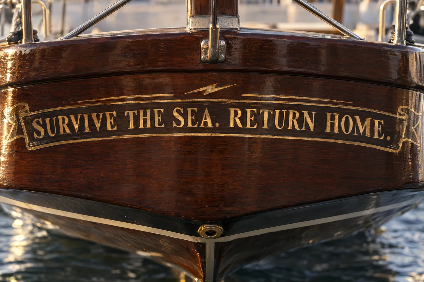 The words Survive the sea. Return home. painted in gold leaf on the foot of a varnished transom